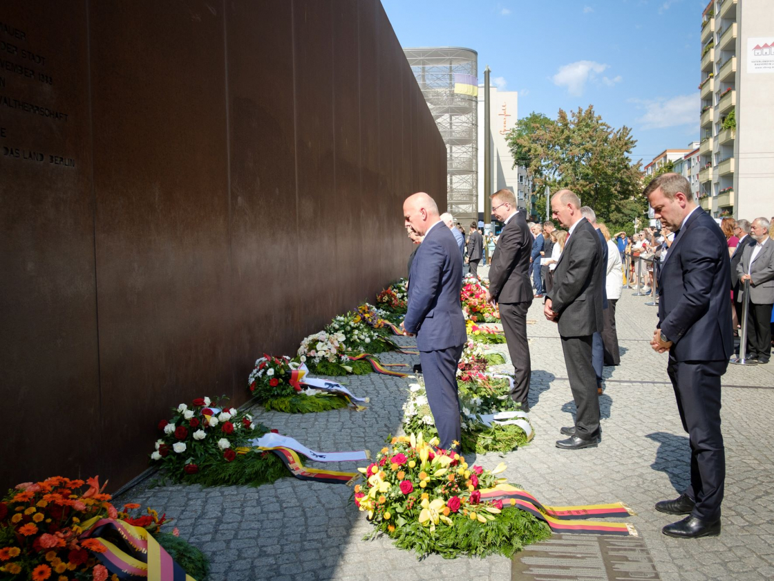 Personen stehen am Denkmal vor Kränzen auf dem Boden