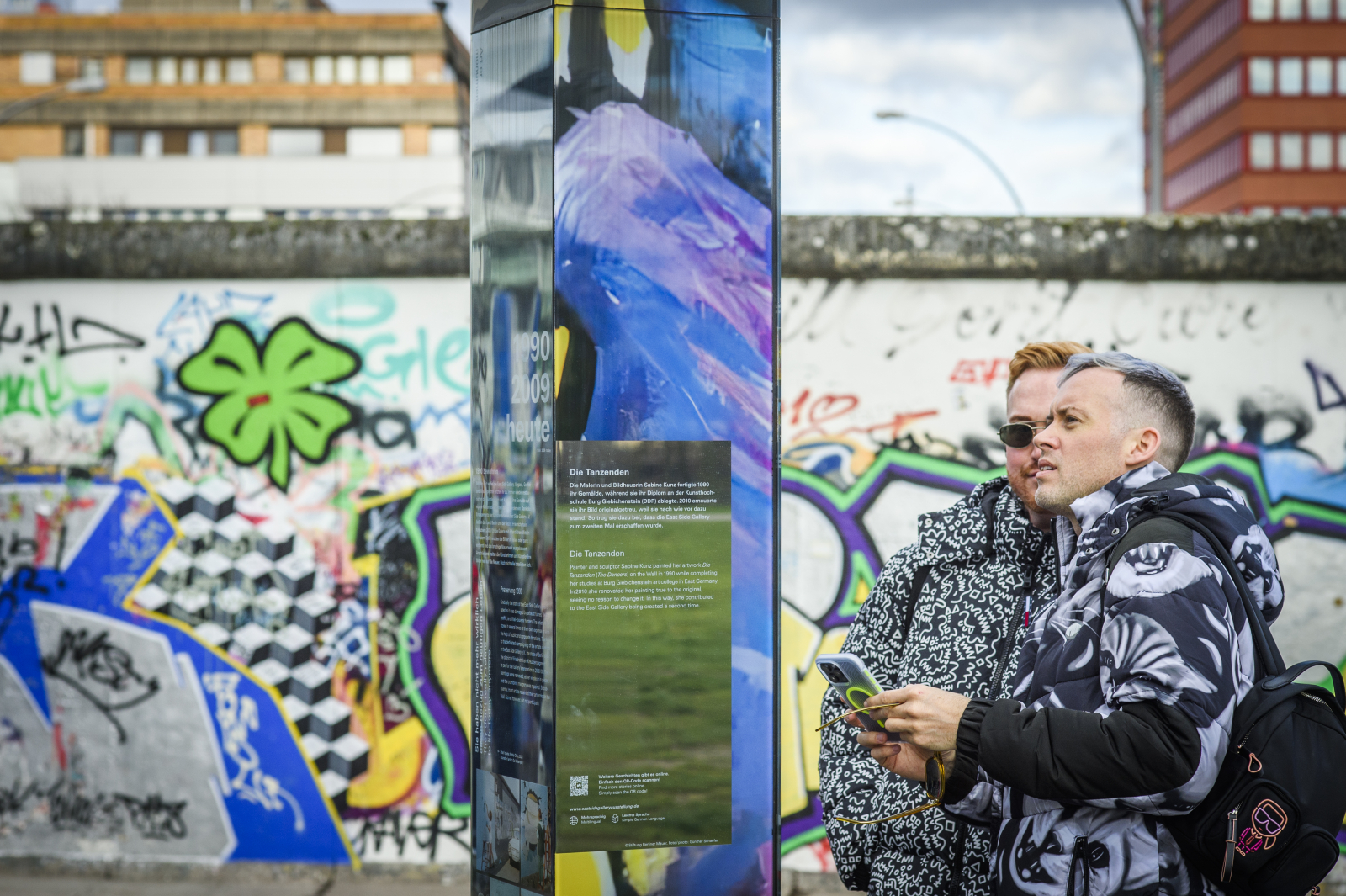Besuchende an einer Infostele. Im Hintergrund die bemalte Mauer.