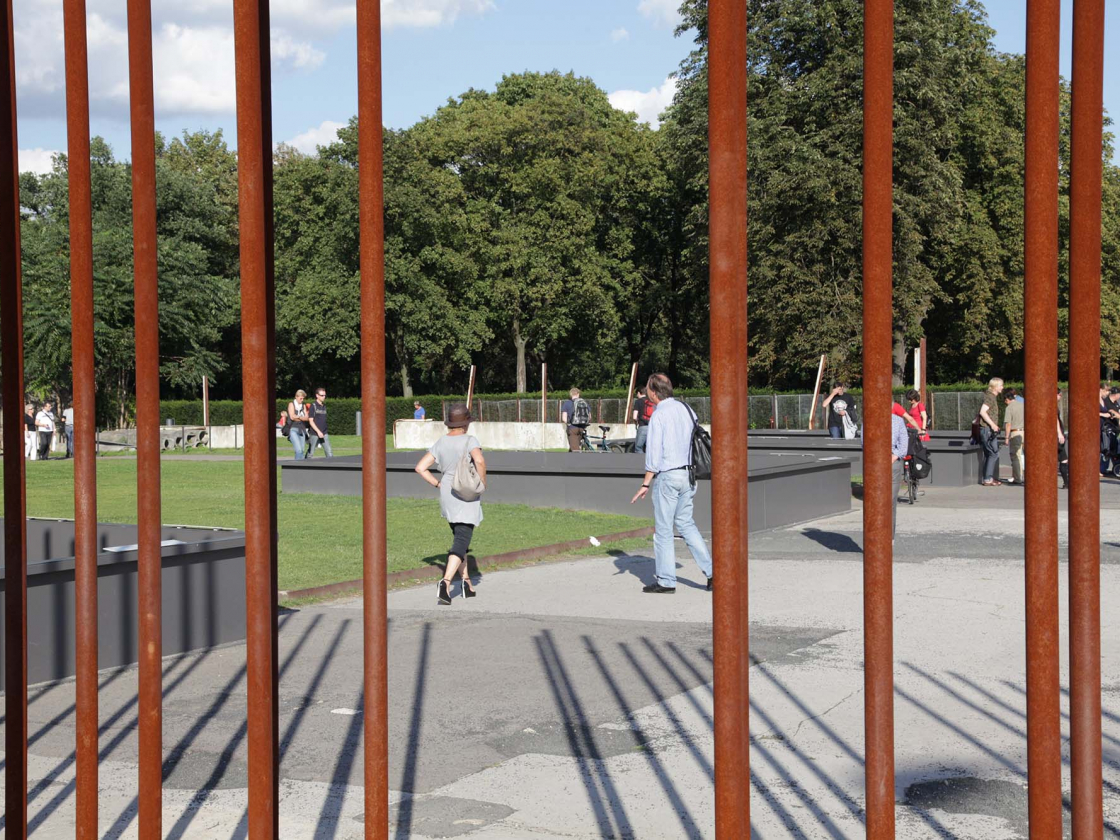 Das Außenareal der Gedenkstätte Berliner Mauer im Sommer. Zu sehen sind vertikale Eisenstäbe, welche den Mauerverlauf kennzeichnen und grüner Rasen.