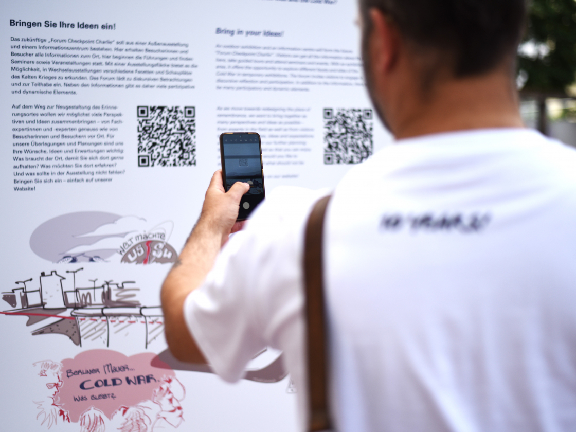 Person scannt einen QR-Code auf einer Infotafel am Checkpoint Charlie