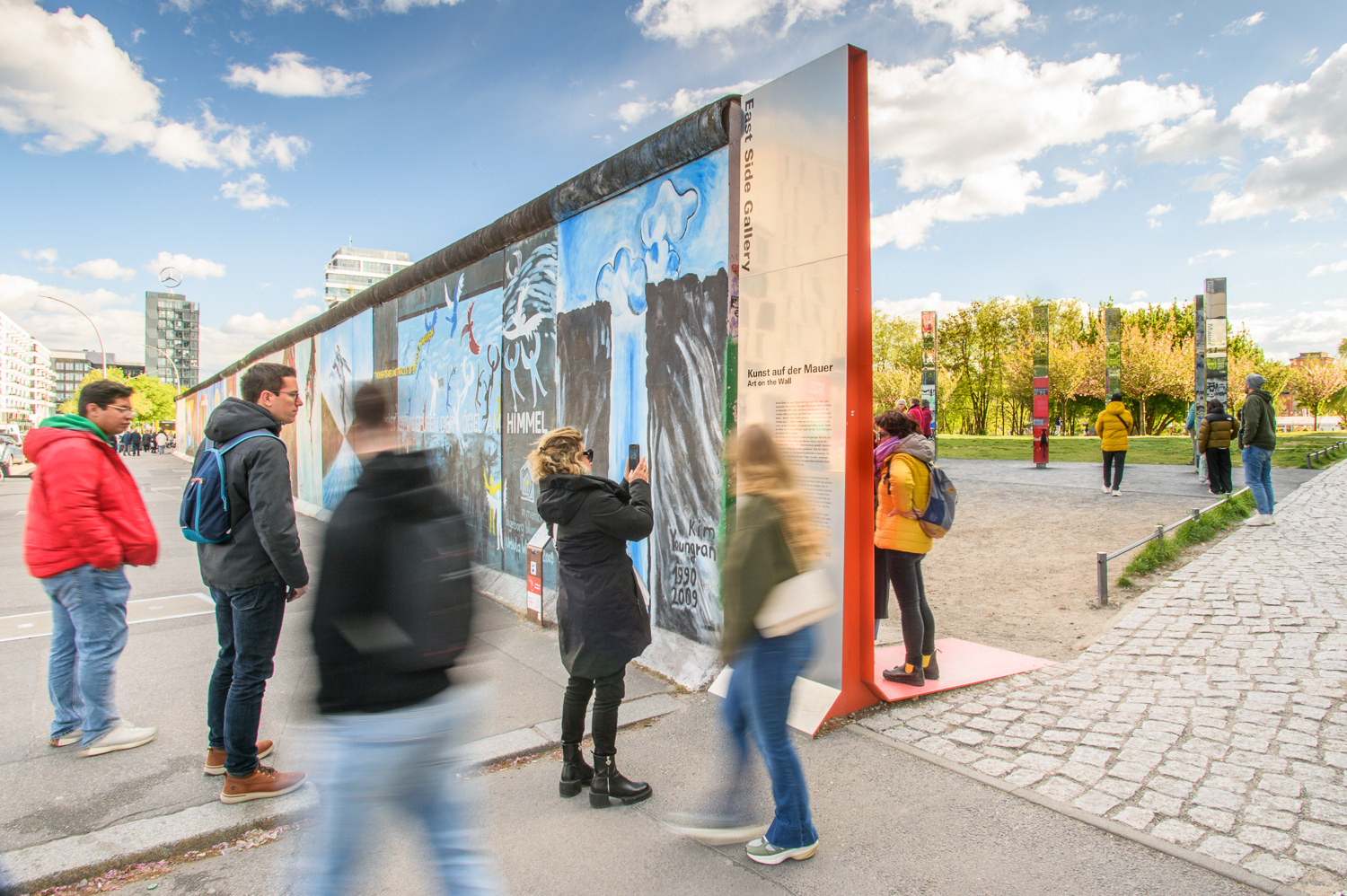 Besuchende an der East Side Gallery. Im Zentrum eine Infotafel in Form eines Mauerwinkels